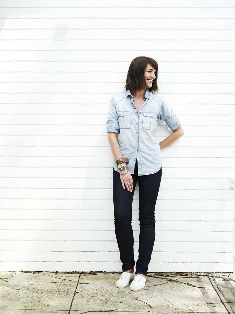 Megan Morton looking sideways and smiling, standing in front of a white brick wall